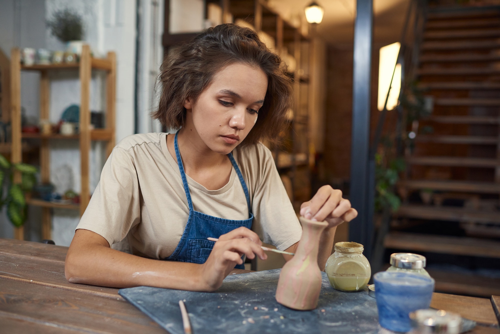 Young female artisan in workwear holding paintbrush and jug